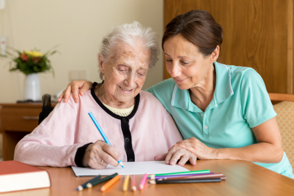 A woman and an older woman sit at a desk. The woman is helping the older woman write in a notebook.