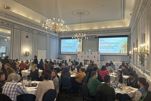 People sit around round dining tables in a brightly lit room with two large screens sowing a presentation