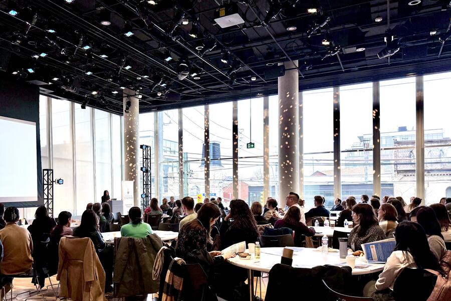 The audience watching as Dr. Shaheen Darani welcomes them to DWED 2025 in a large auditorium with floor-to-ceiling windows.