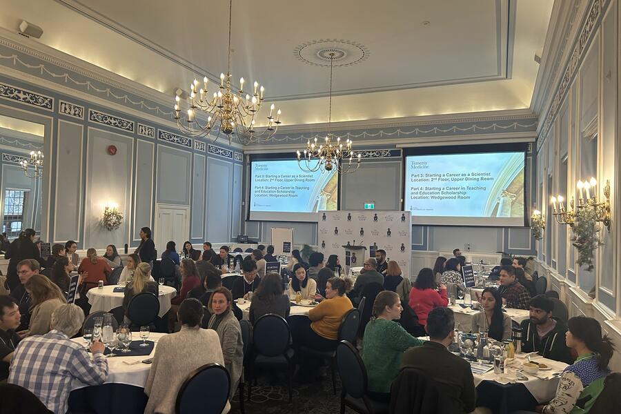 People sit around round dining tables in a brightly lit room with two large screens sowing a presentation