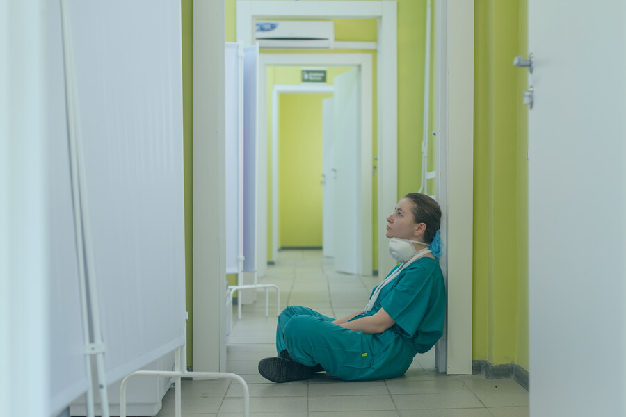 A health care worker in scrubs sits on the floor in a hospital corridor, looking exhausted