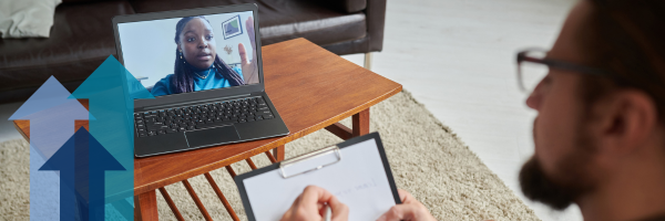 A man takes notes while in conversation with a women on a video call. The woman is visible on the screen of a laptop sitting on a coffee table.