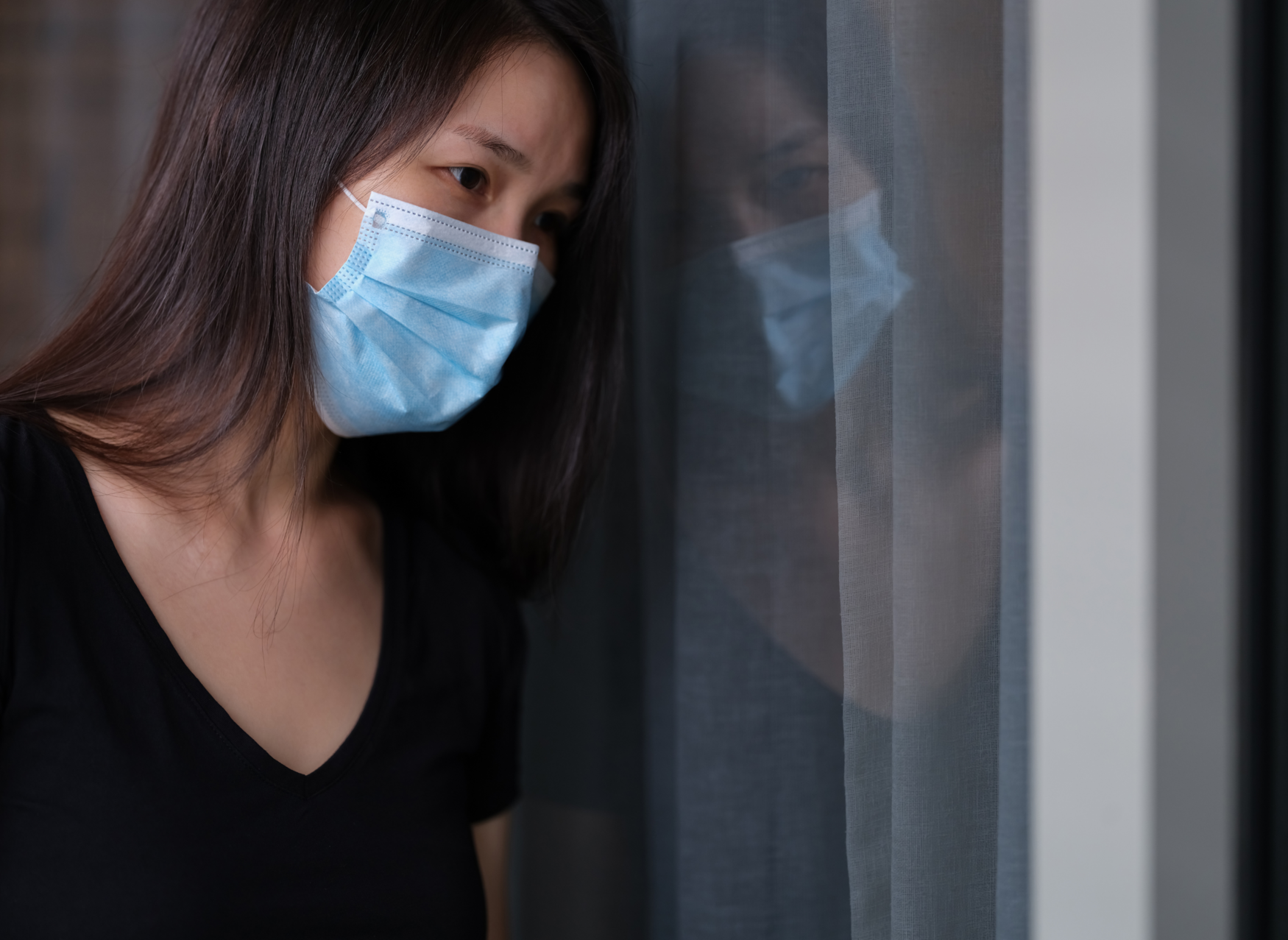 A woman leans against a window while wearing a medical mask. Her expression is tired and sad.