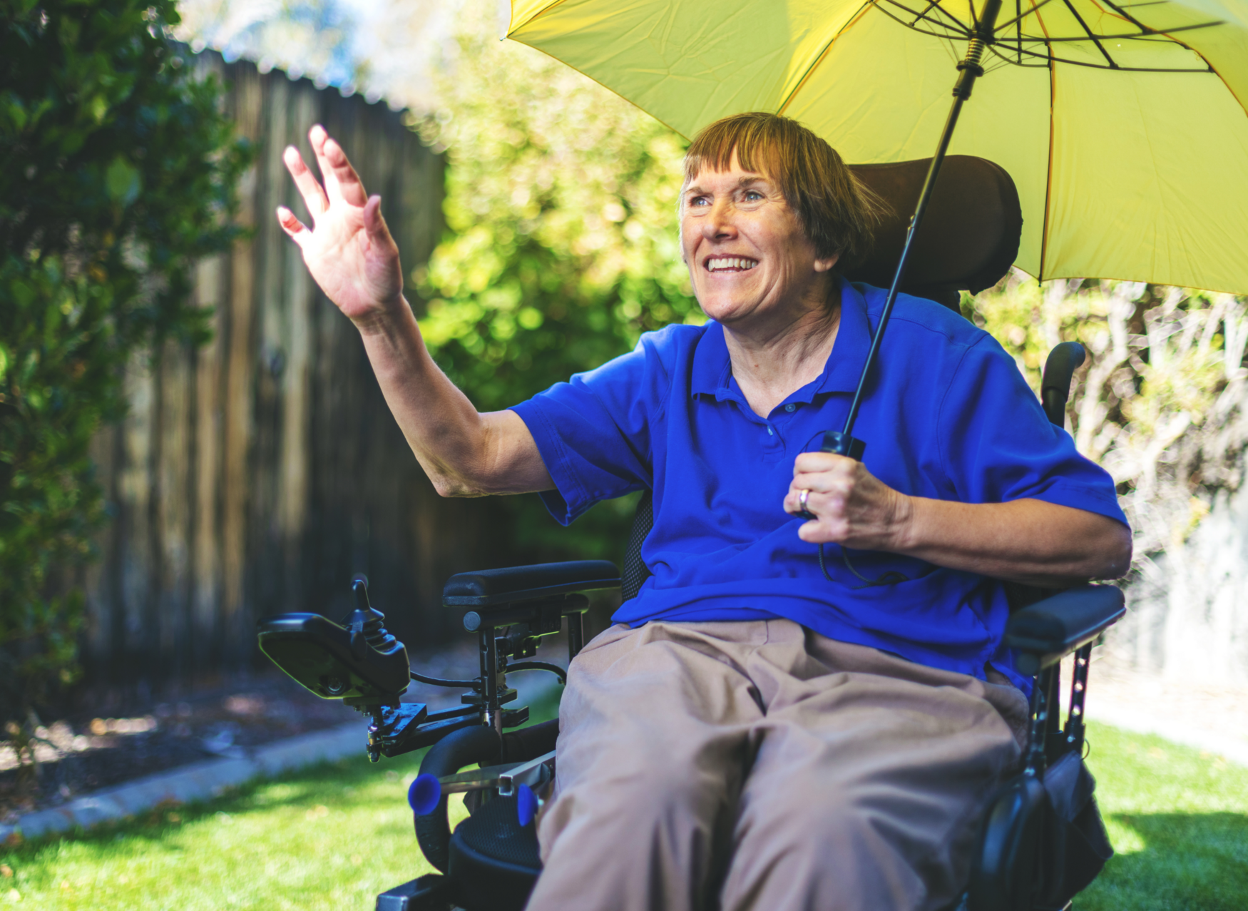 A middle-aged woman in an electric wheelchair sits outside. She is holding a yellow umbrella, smiling, and waving at someone who is not in the photo.