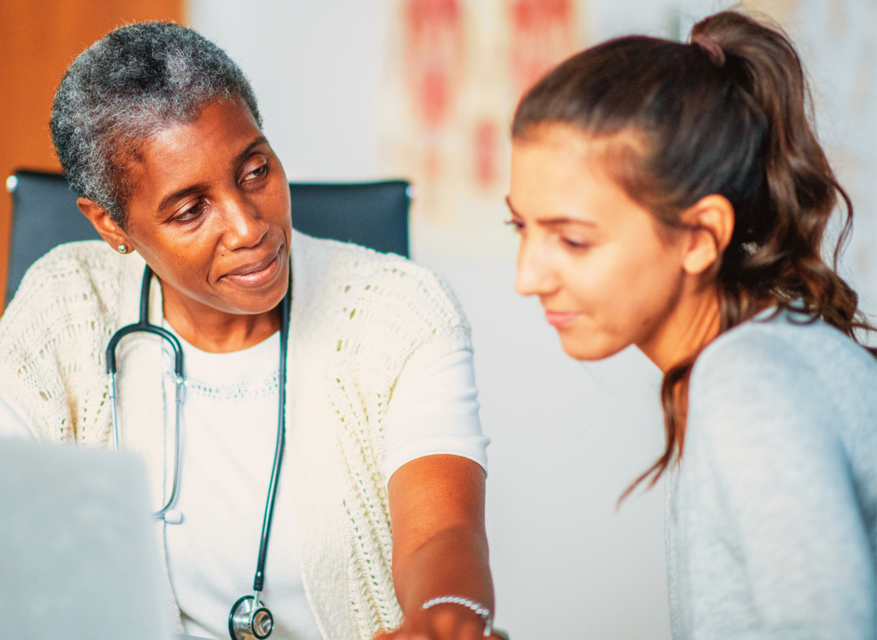 An older woman wearing a stethoscope sits with a younger woman in a medical office, looking at a laptop together.