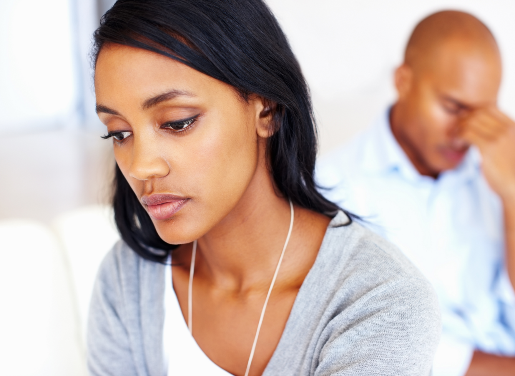 A sad woman sits with her head bowed, while her frustrated partner sits with a hand to his face behind her