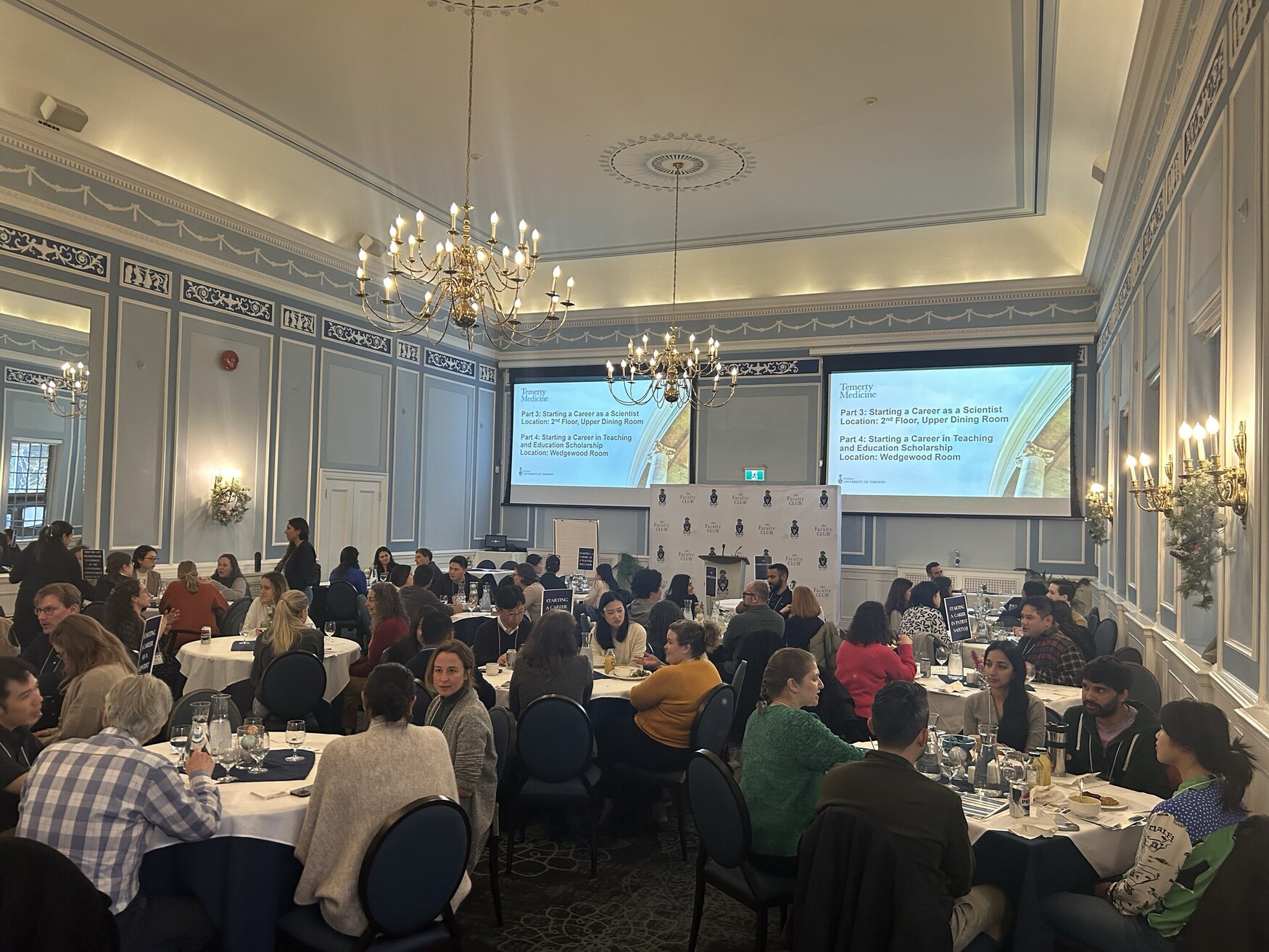 People sit around round dining tables in a brightly lit room with two large screens sowing a presentation