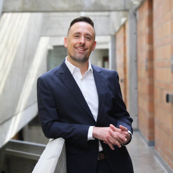 Anthony Ruocco, a middle aged man wearing a suit, smiles at the camera as he stands in a modern, brick and glass corridor.