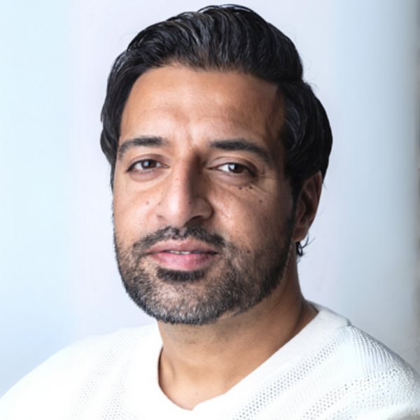 A middle-aged man with a beard and neat, dark hair looks at the viewer. He is wearing a white t-shirt and a grey background is behind him.