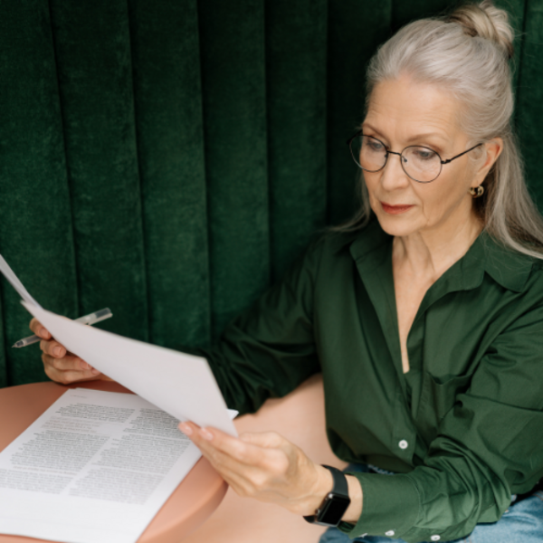 An older woman sitting at a small table thoughtfully reading a document.